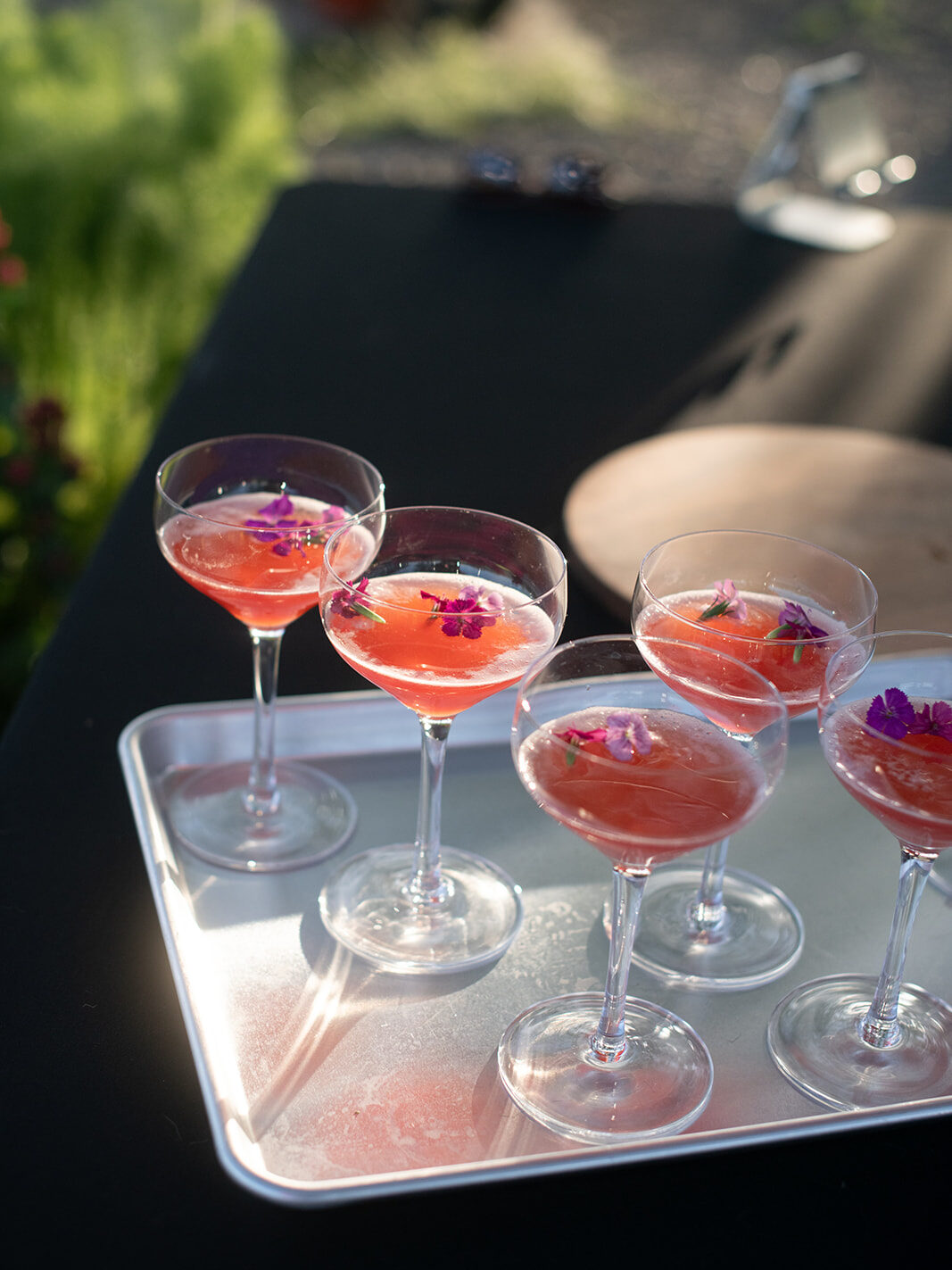 Side-view of seven bright pink non-alcoholic cocktails on a silver tray, resting on a black tablecloth, with sunlight shining through.