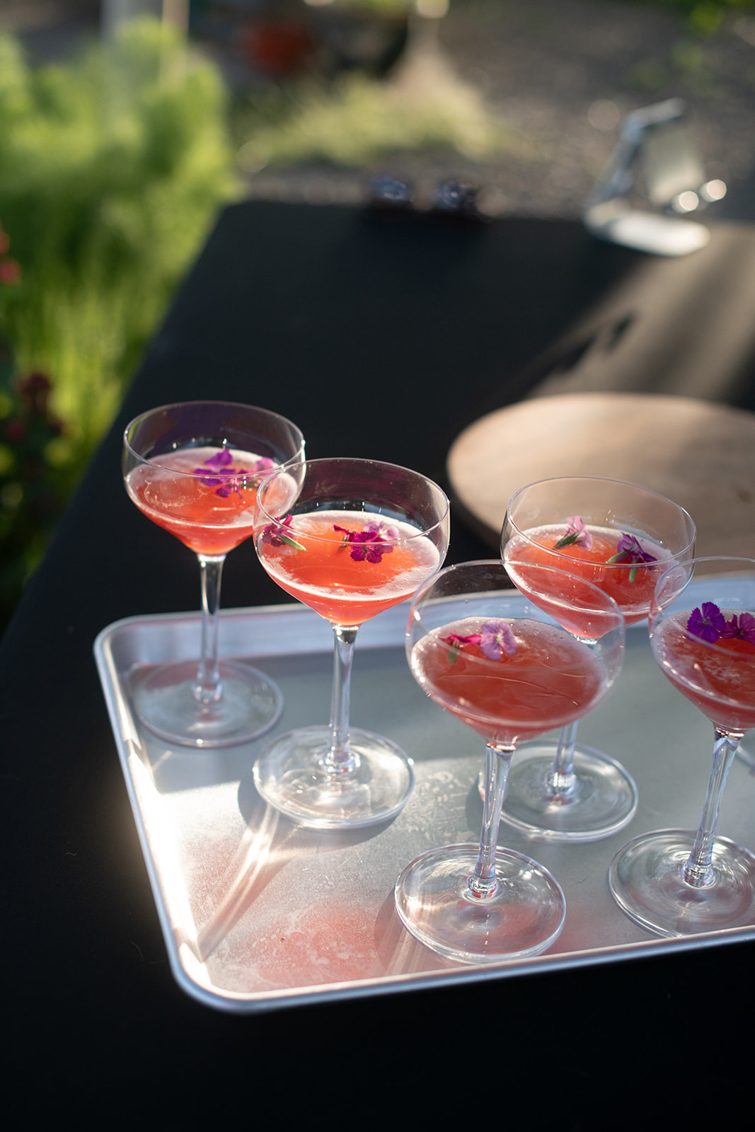 Side-view of seven bright pink non-alcoholic cocktails on a silver tray, resting on a black tablecloth, with sunlight shining through.