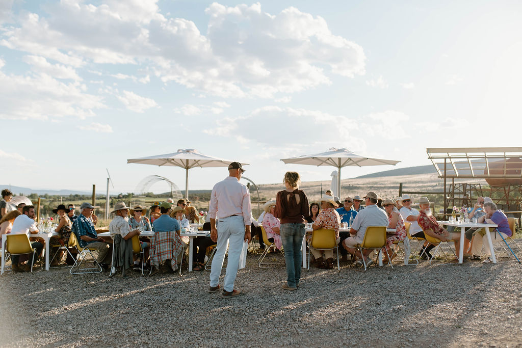 Two winemakers addressing a group of people at an outdoor wine-paired dinner in the summertime.