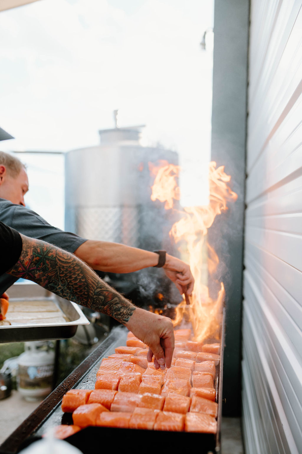 Two chefs cooking 45 pieces of salmon on a grill with flames.